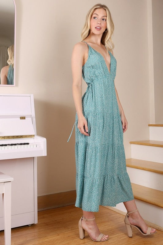 A woman with long blonde hair stands against a neutral background wearing the SL V neck tier dress, which features a teal color and polka dot pattern.