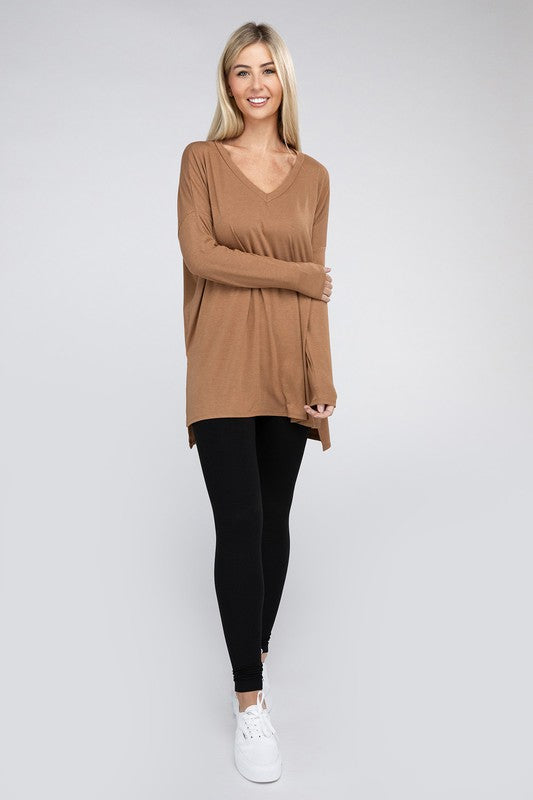 A woman with long, blonde hair is wearing a loose-fitting, brown Dolman Long Sleeve V-Neck Side Slit Hi-Low Hem Top and black pants. She poses with one hand on her hip and a neutral facial expression against a light grey background.