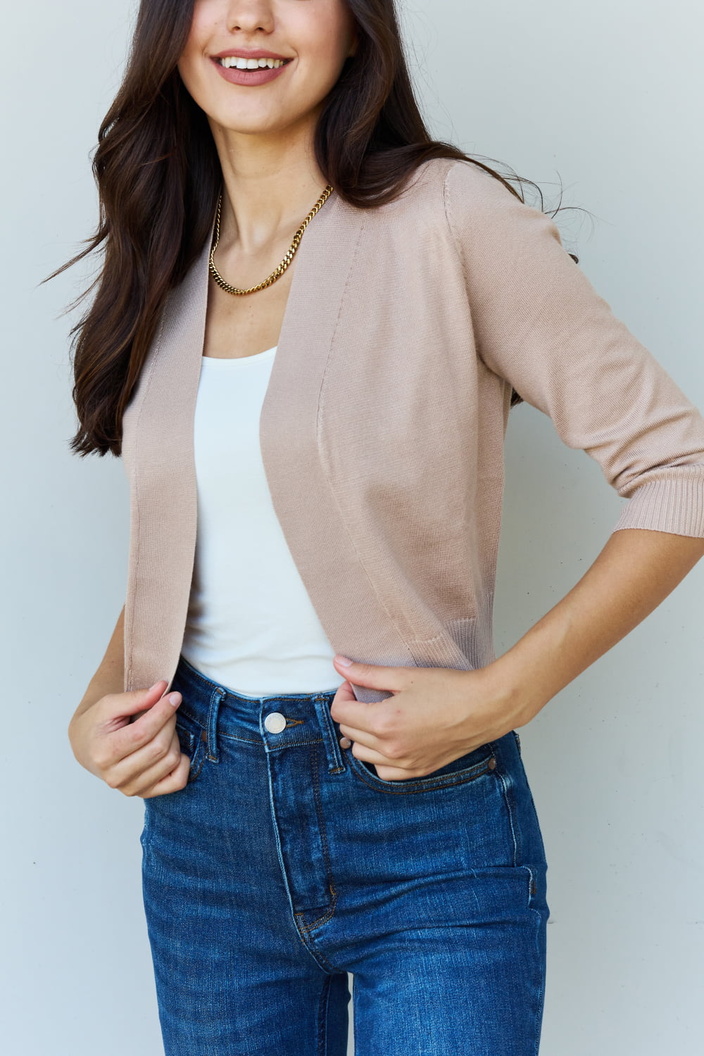 Woman with long dark hair wearing the versatile Doublju My Favorite Full Size 3/4 Sleeve Cropped Cardigan in Khaki over a white top and blue jeans, standing against a plain background.