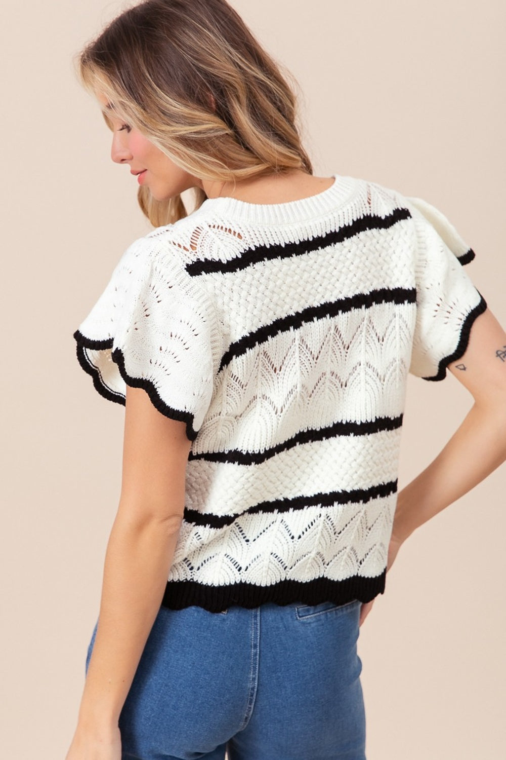 A woman with long blonde hair wearing a BiBi Pointelle Contrast Striped Short Sleeve Knit Top and white shorts smiles in a brightly lit room. A colorful painting is visible in the background, adding a touch of vibrancy to her sophisticated, stylish wardrobe.