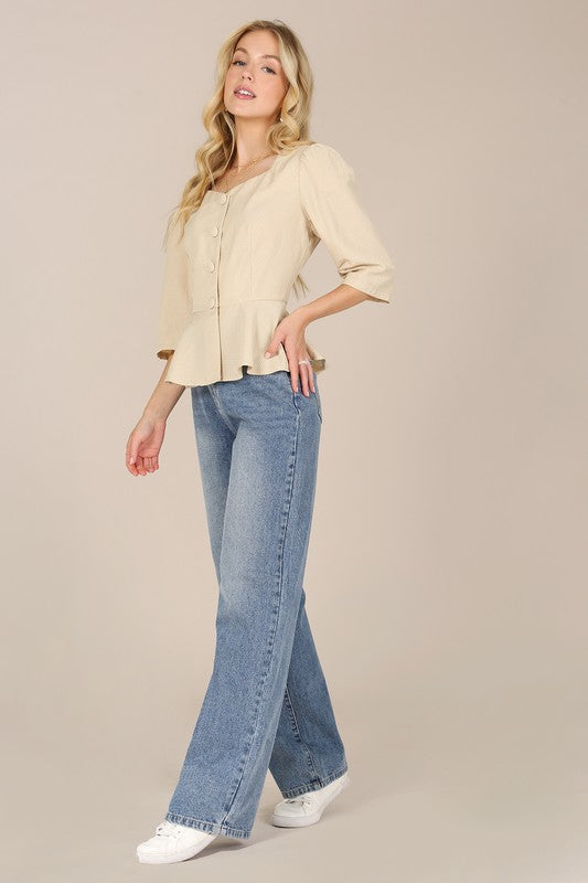 A person with long blonde hair is posed against a plain background, wearing a beige 3/4 sleeve front button blouse paired with puff sleeves and blue jeans.