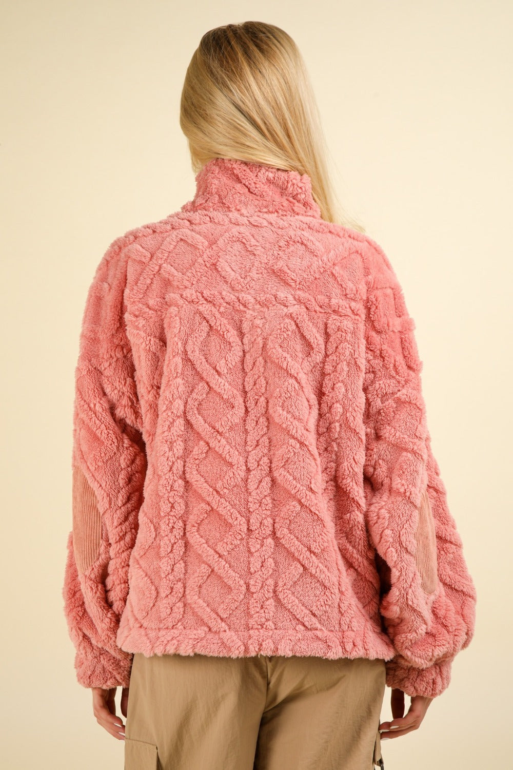 A person with long blonde hair wears a VERY J Fuzzy Fleece Half Zip Cable Pattern Sweatshirt in pink, paired with beige pants, standing against a plain light background. The sweatshirt is adorned with textured details and elbow corduroy patches.