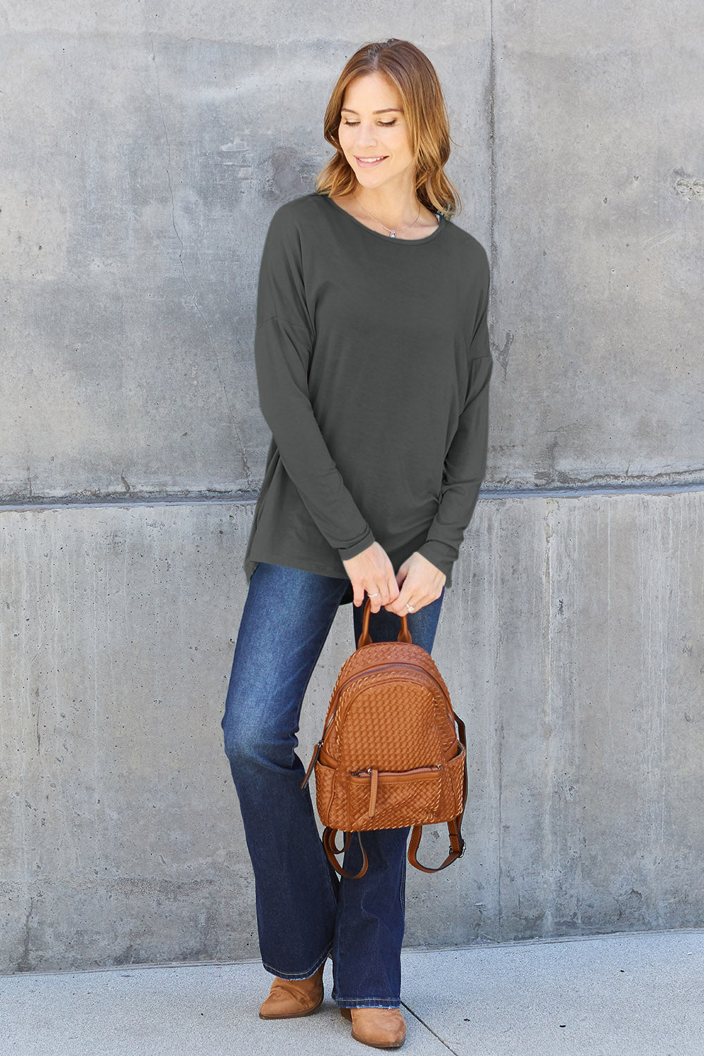 A woman with shoulder-length hair wearing a Basic Bae Full Size Round Neck Dropped Shoulder T-Shirt in green and a pair of jeans, exuding a basic style, stands against a concrete wall. She's holding an imported brown woven backpack on her shoulder while touching her hair with her hand.