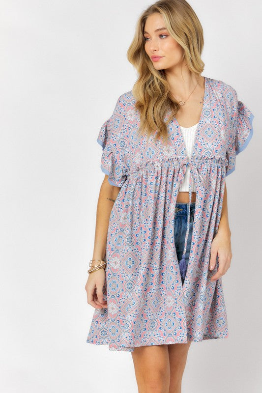 A woman stands against a white background wearing the Printed Short Sleeve Ruffle Kimono over a white top and denim shorts, effortlessly blending style and comfort.