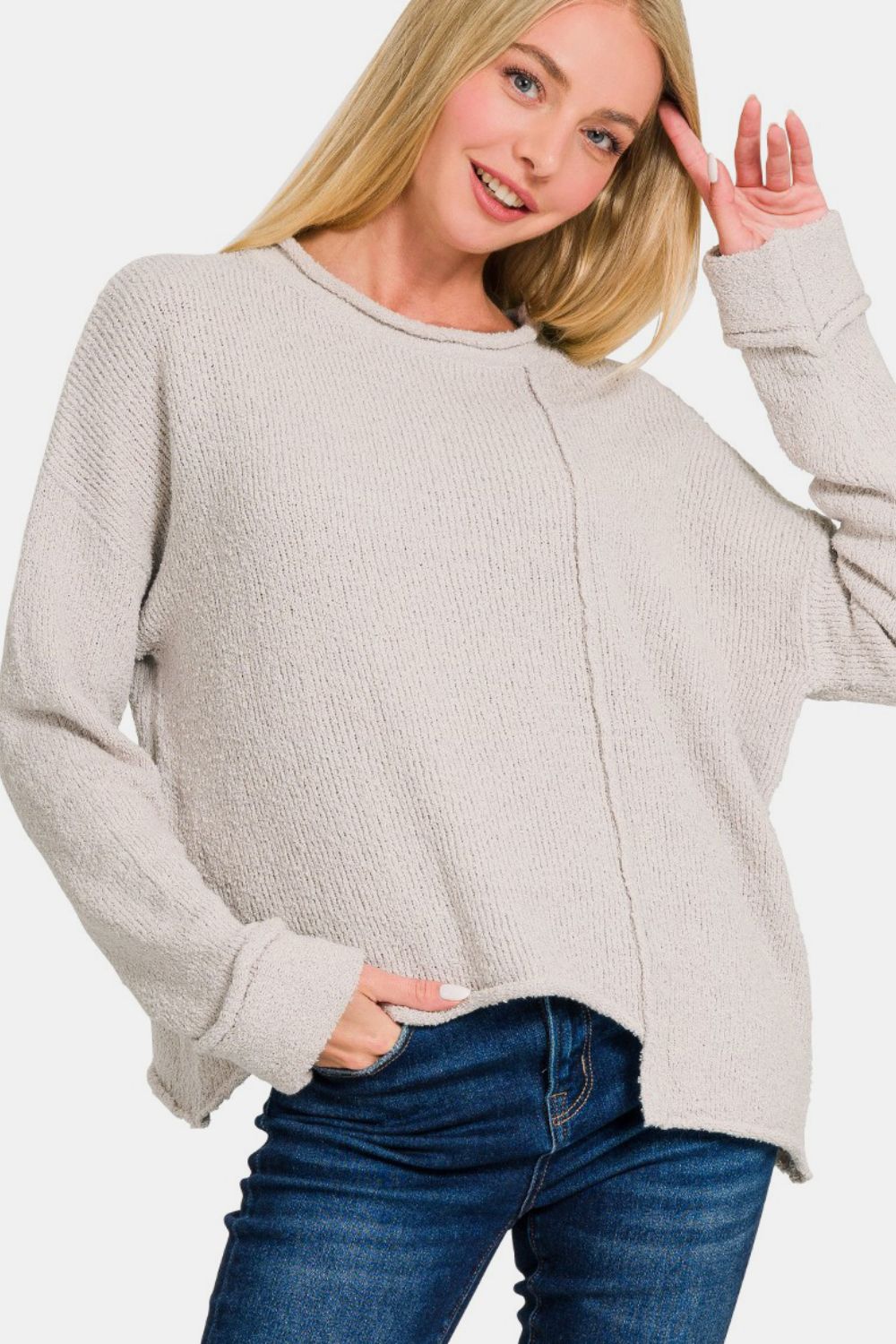 A woman in a light beige Zenana Asymmetric Hem Drop Shoulder Sweater poses with a hand near her head and another in her pocket, smiling slightly. She is wearing blue jeans against a plain white background.