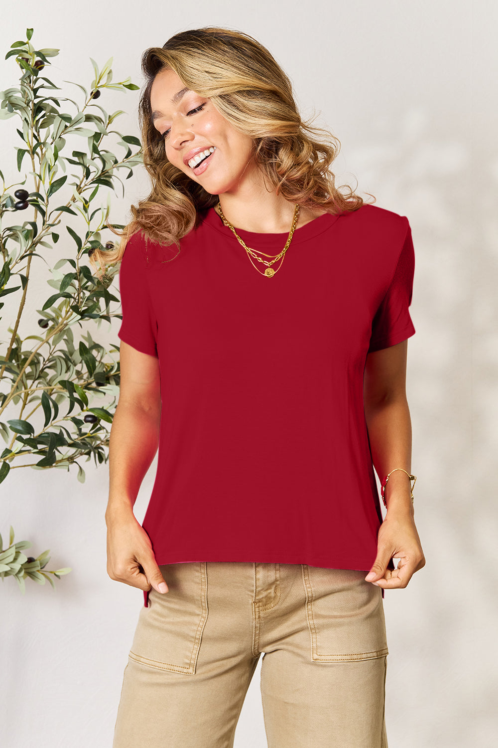 A person with wavy hair, wearing the Basic Bae Full Size Round Neck Short Sleeve T-Shirt in bright pink and beige pants, stands near a plant, smiling and looking to the side. The outfit embodies a basic style and offers slightly stretchy comfort, ideal for those who value effortless elegance.