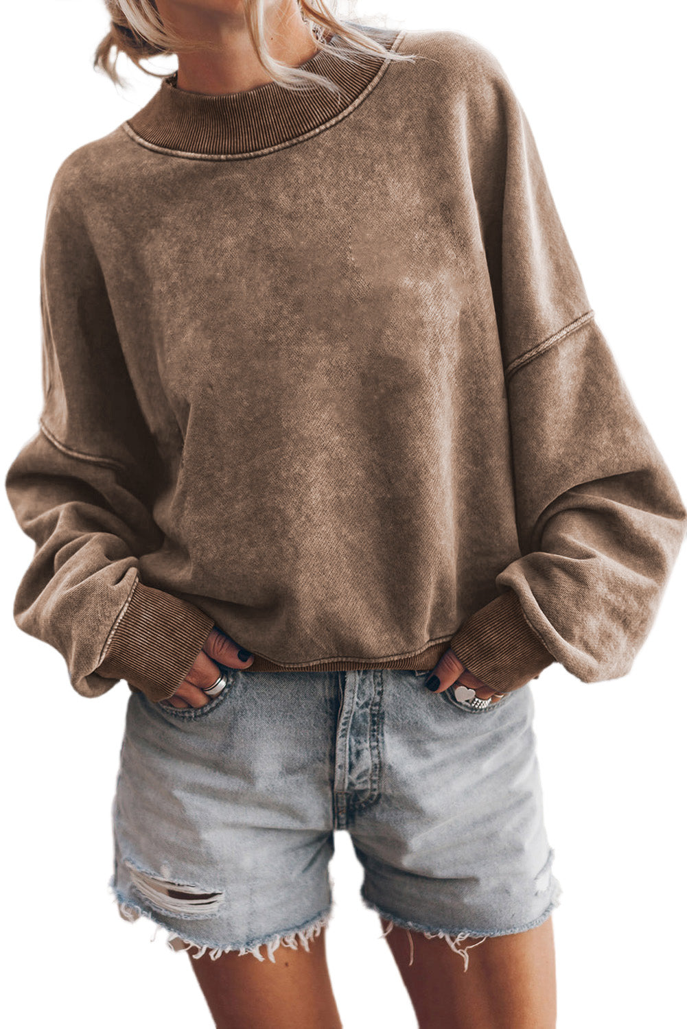 A person is seen wearing a brown Drop Shoulder Crew Neck Pullover Sweatshirt with rolled-up sleeves, paired with light blue jeans and standing with one hand in their pocket. Multiple bracelets are visible on their wrist while the background appears blurred.