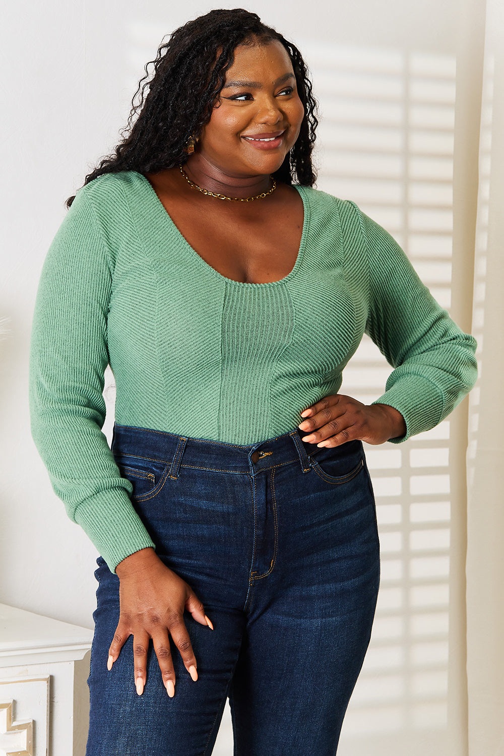 A person wearing a fashion-forward Basic Bae Scoop Neck Long Sleeve Top in green and dark jeans stands indoors, smiling and looking to the side.