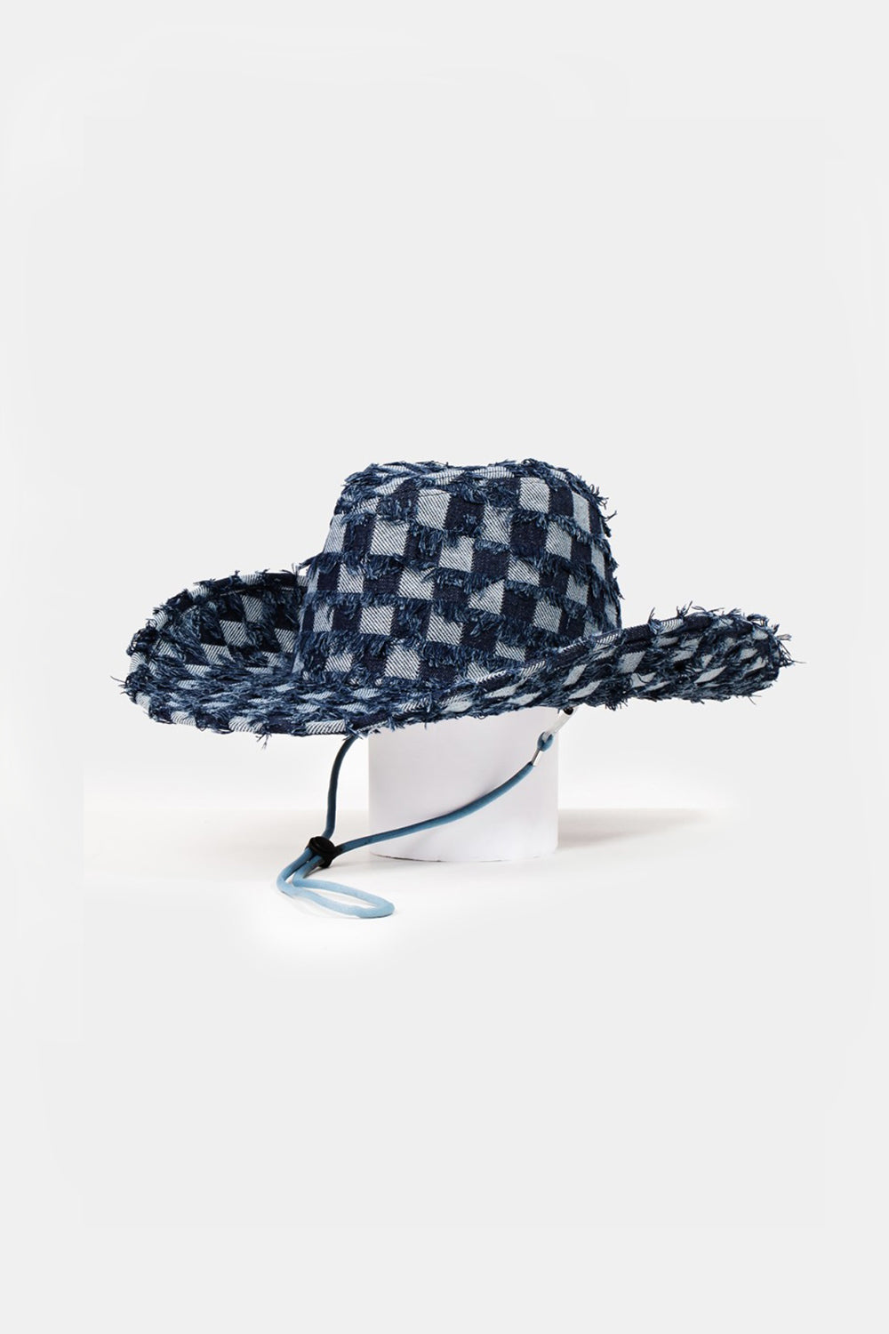 The Fame Checkered Fringe Denim Cowboy Hat, featuring a wide brim and adorned with a blue checkered pattern along with frayed denim edges, rests on a pristine white surface. It includes an adjustable inner string and a matching blue chin strap for added style and functionality.