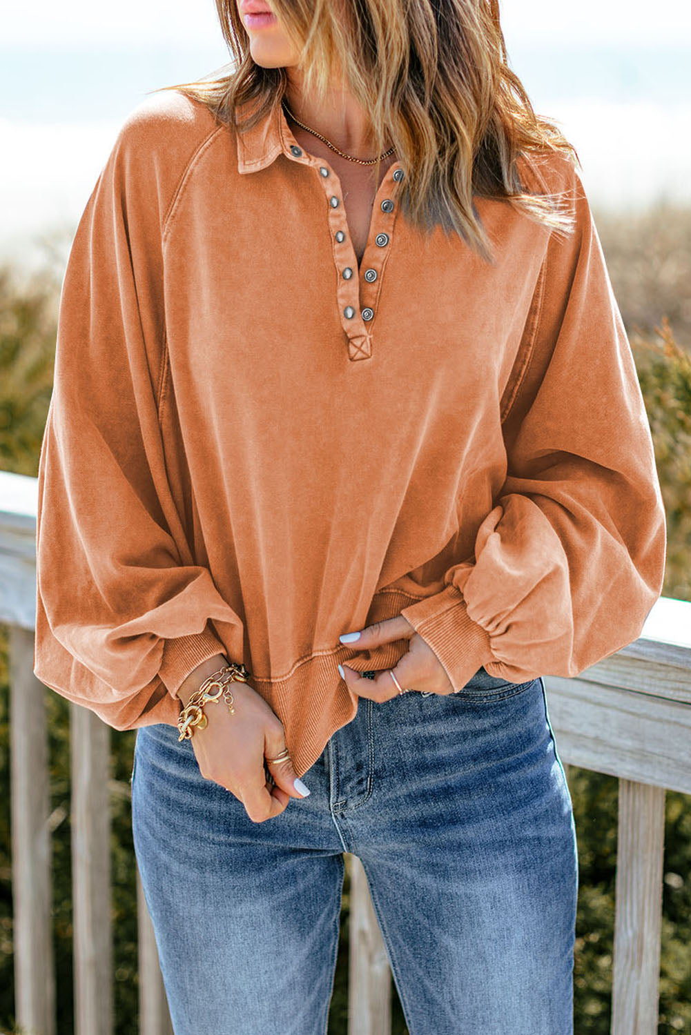 A person wearing an orange washed snap buttons lantern sleeve pullover sweatshirt and blue jeans stands with one hand on their hip, facing away from the camera, leaning slightly on a wooden rail outdoors.