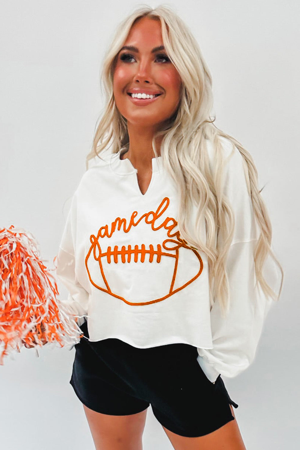 A woman with long blonde hair dressed in a chic White Game Day Lettering Rugby Football Notched Neck Sweatshirt and blue jeans stands near a Christmas tree adorned with red and white ornaments and ribbon.