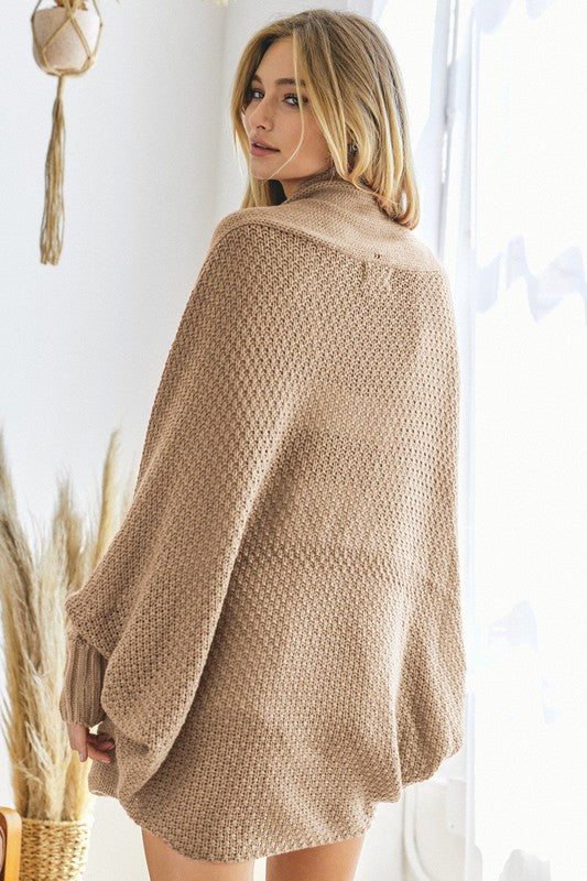 A person stands against a plain background, wearing a Pattern Knit Dolman Sleeve Solid Slouch Cardigan in textured blue over a white sweater with distressed white shorts, hands in cardigan pockets.