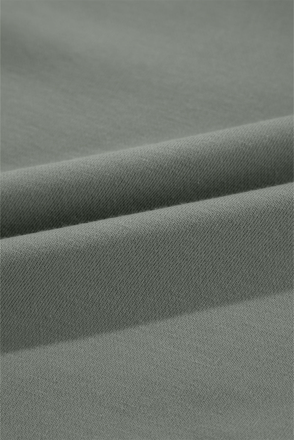 A woman with wavy blonde hair is seen from behind, wearing a grayish-green round neck sweatshirt with patchwork sleeves and blue jeans. She carries a brown shoulder bag against a backdrop of woven wall decorations.