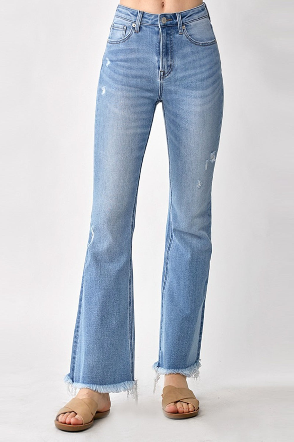 A person wearing RISEN High Rise Frayed Hem Bootcut Jeans in light-blue stretchy denim paired with tan sandals, standing against a plain background.