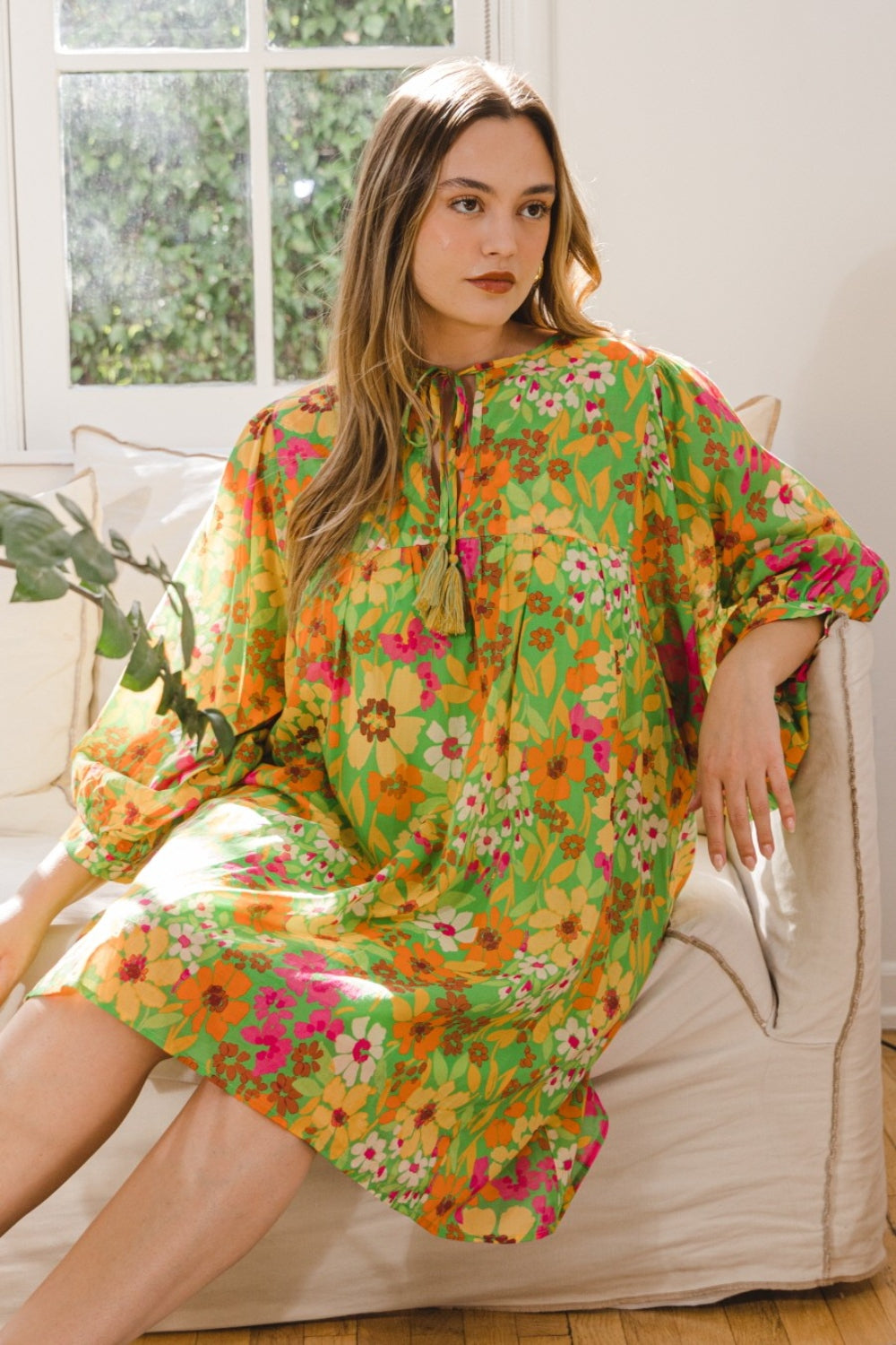 A person is seated on a beige couch, adorned in an ODDI Full Size Floral Tied Neck Mini Dress, beside a window that displays the lush greenery outside.