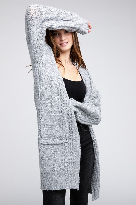 A woman wearing a white tube top, distressed denim shorts, a beige hat, and the Twist Knitted Open Front Cardigan With Pockets poses against a plain background.