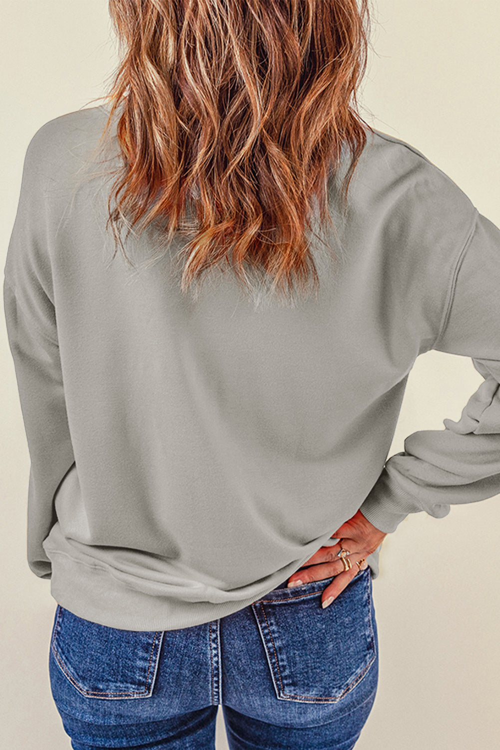 Person wearing a Gray Plain Crew Neck Pullover Sweatshirt and blue jeans with a hand in their pocket, adorned with layered necklaces.