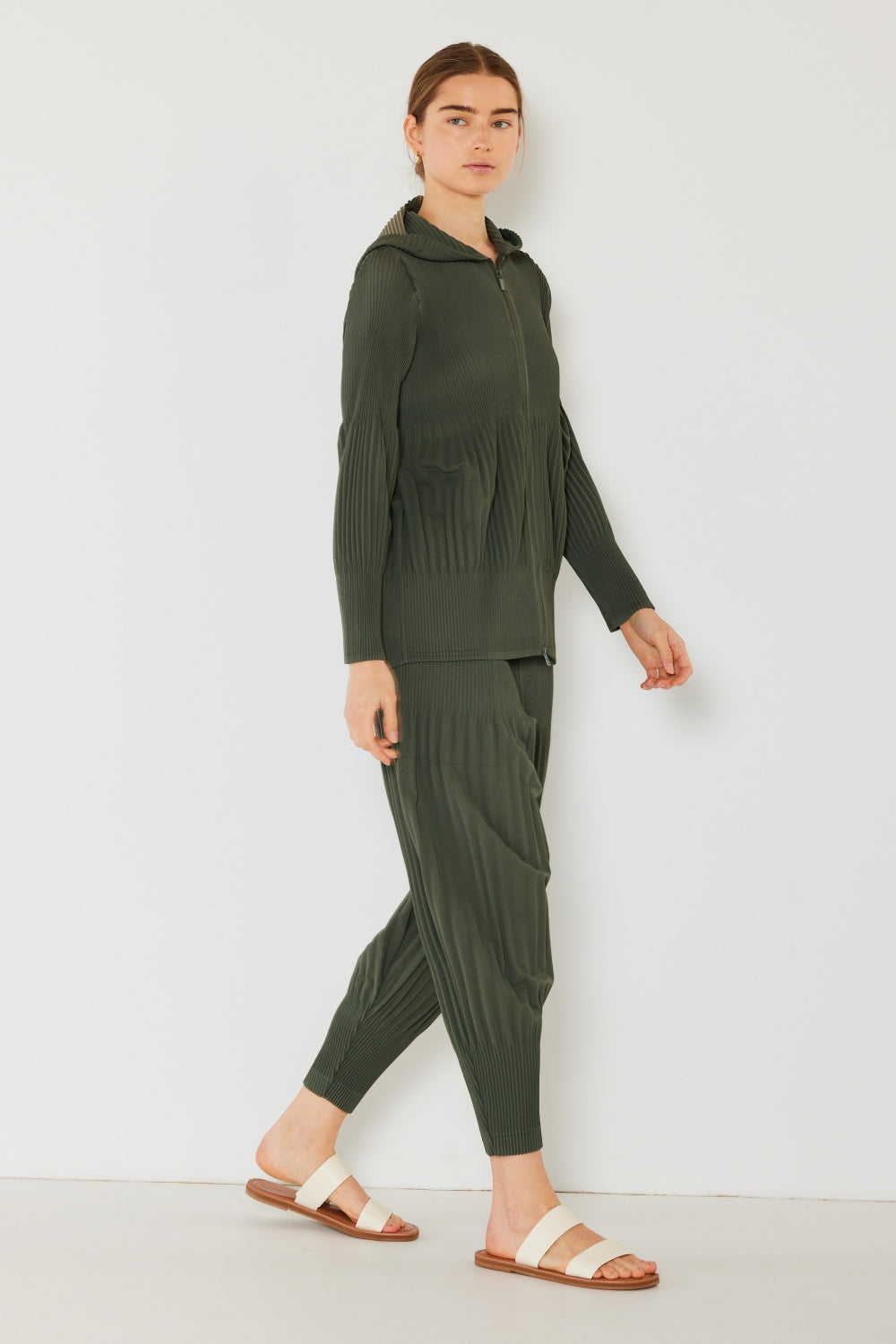 A person stands against a plain background, wearing a matching dark green hooded top and Marina West Swim Pleated Unisex Aladdin Plisse Pants with a ribbed texture, complemented by white sandals.