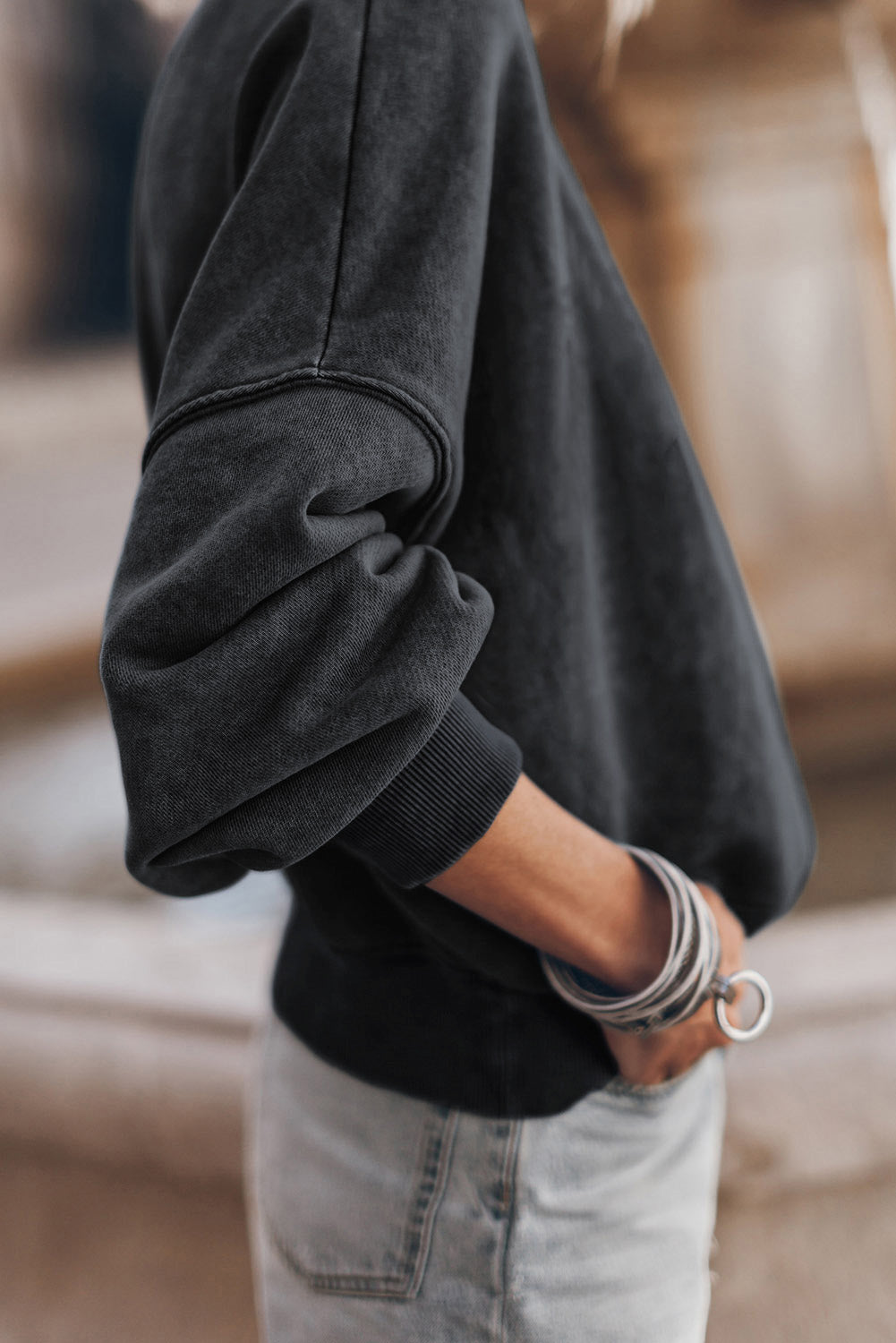 Person wearing a black drop shoulder crew neck pullover sweatshirt with rolled-up sleeves and light blue jeans, standing with one hand in their pocket, showing multiple bracelets on their wrist.