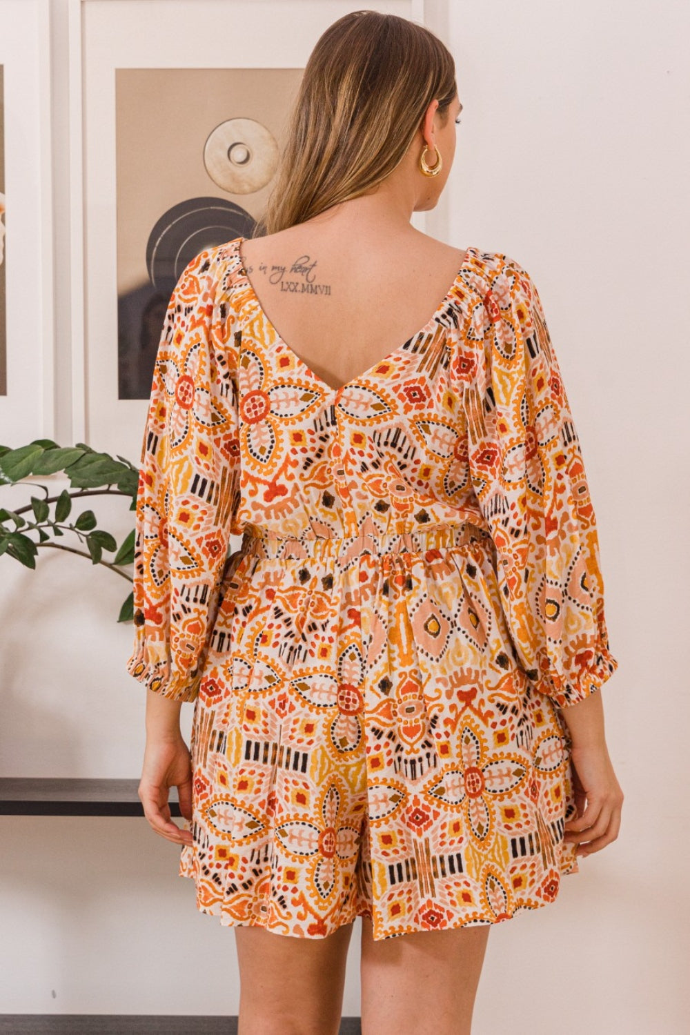 A person stands wearing the ODDI Full Size Printed Ruff Sleeve Romper with Pockets, featuring vibrant orange, red, and yellow designs. They are positioned in front of a wall adorned with framed art and a plant.