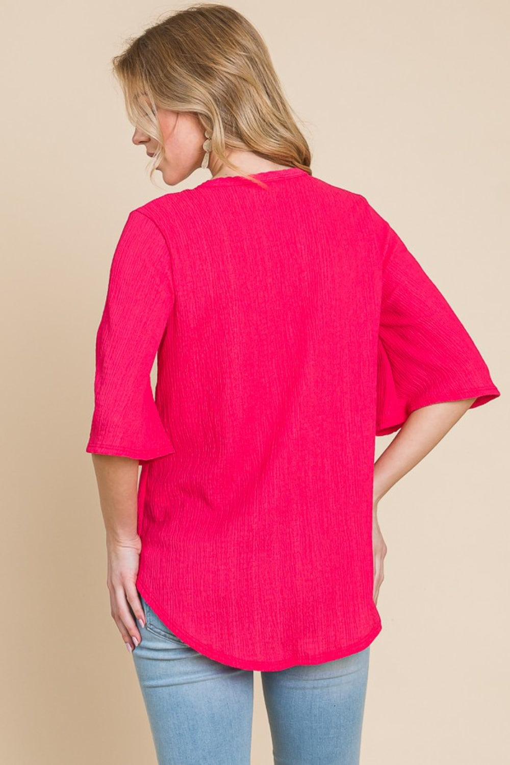 A person is wearing the BOMBOM Texture Decorative Button V-Neck Top, a chic design in bright pink featuring elegant details like short bell sleeves, and pairing it with blue jeans, stands against a beige background.