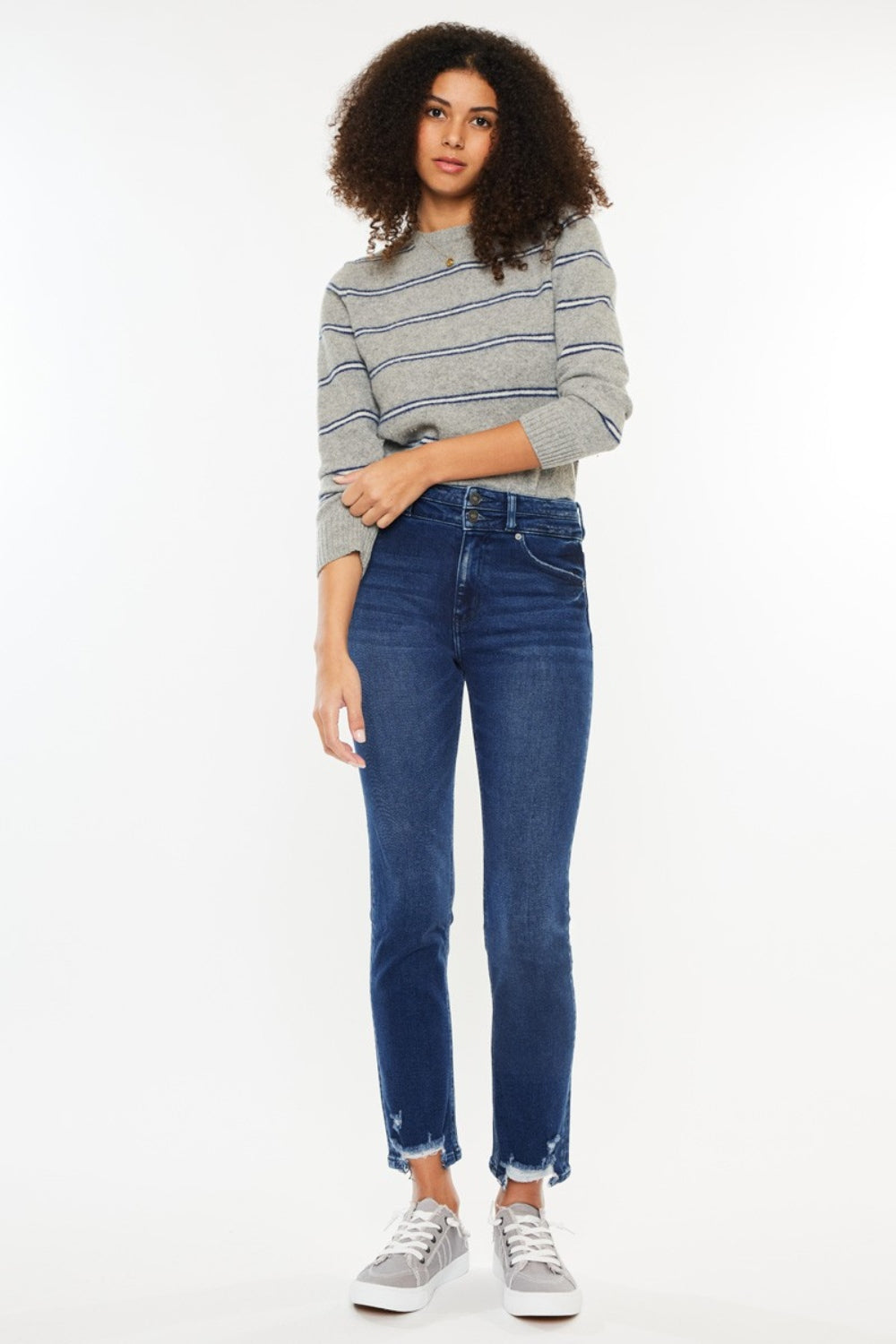 A person with curly hair is wearing a gray striped sweater, Kancan High Rise Slim Straight Jeans in quality denim, and gray sneakers, standing against a plain white background.