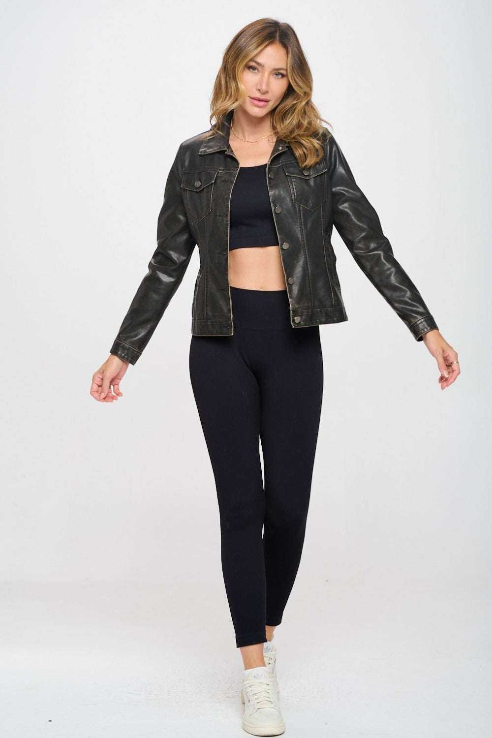 A woman wearing the Coalition LA Button Down Cargo Vegan Leather Shacket over a black crop top and black leggings poses against a white background.