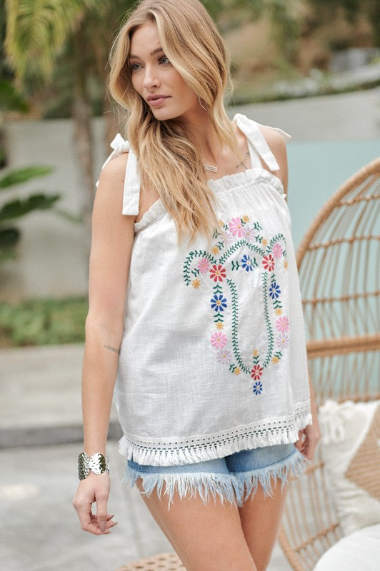 A person wearing an Embroidered Sleeveless Ruffle Tank Top and denim shorts stands outdoors near a wicker chair, adjusting their hair.