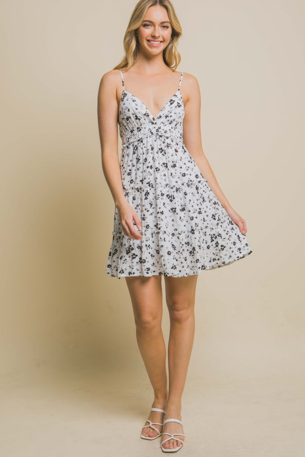 A woman wearing the Love Tree Floral Tiered Cami Mini Dress stands against a beige background, with one hand on her hip and a neutral expression.