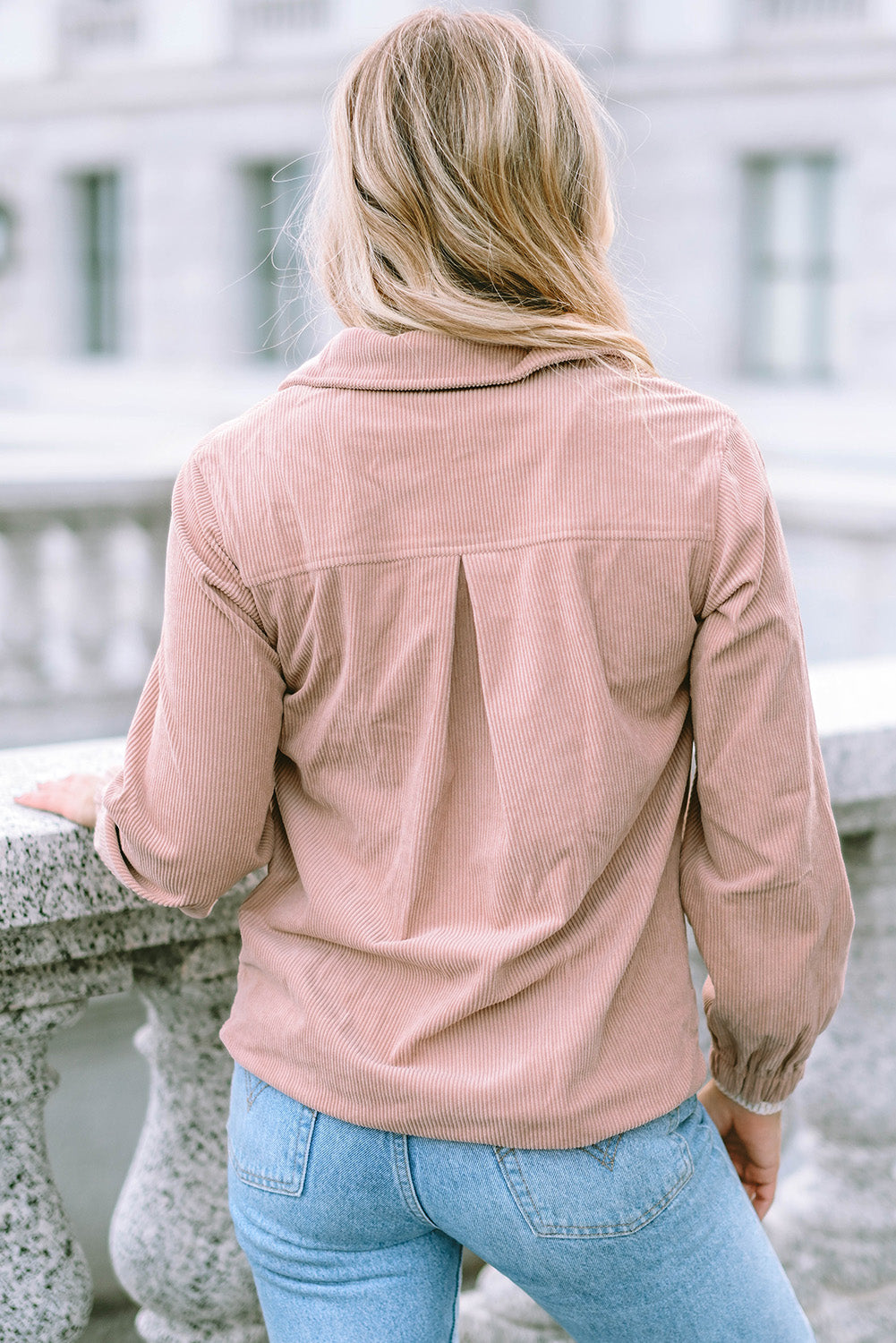 A person with long blonde hair, wearing a trendy Apricot Ribbed Texture Half Zip Collared Sweatshirt and blue jeans, stands facing away and looks over a stone balustrade.