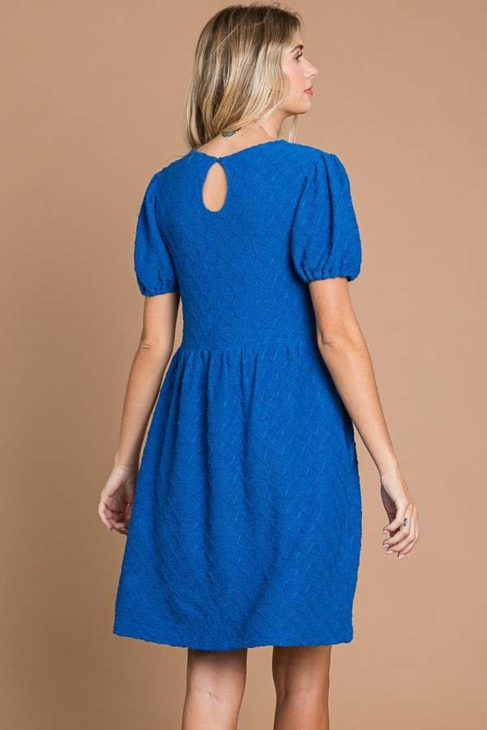 Against a plain brown backdrop, an individual is styled in a Culture Code Texture Round Neck Short Sleeve Dress with Pockets, exuding elegance and flair with its blue textured fabric and chic short puff sleeves.