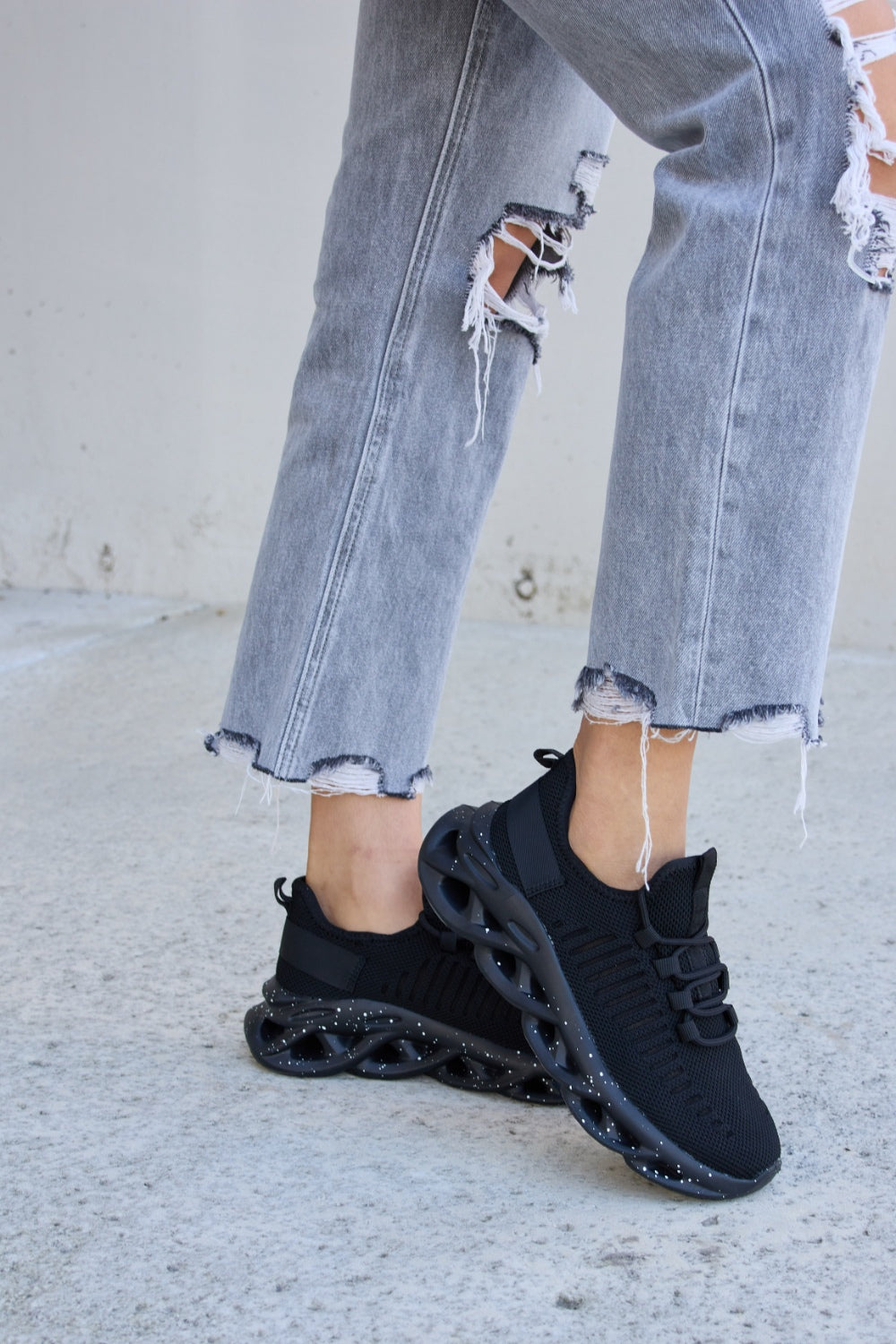 A person wearing ripped gray jeans and Forever Link Mesh Breathable Lace-Up Athletic Shoes is standing on a concrete surface.