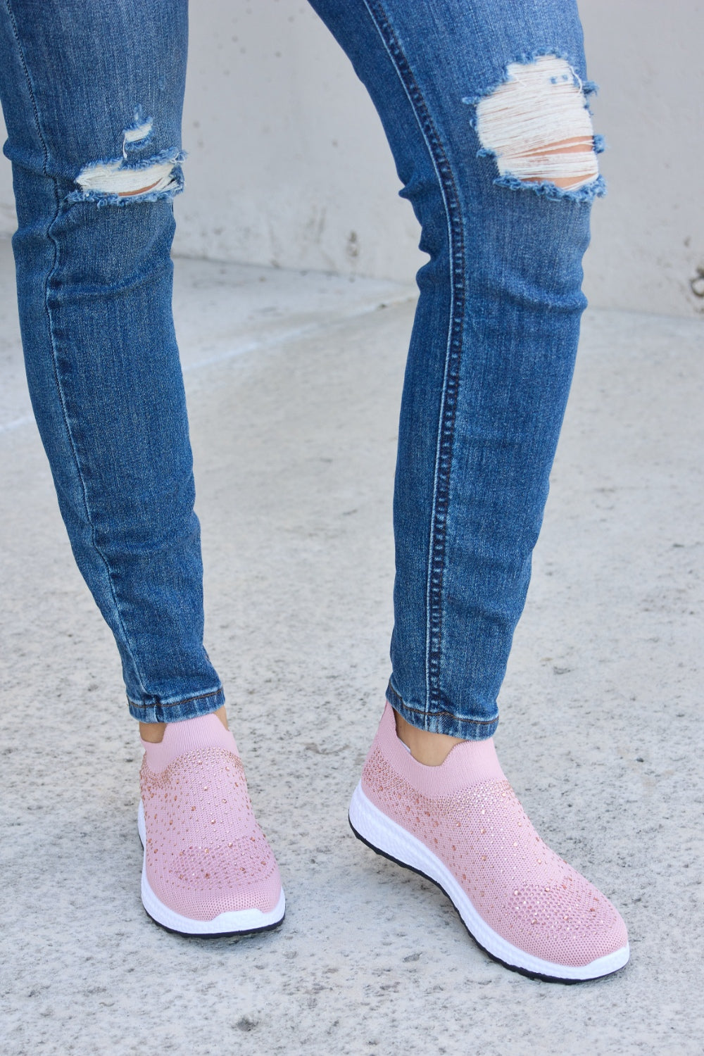 Person wearing Forever Link Rhinestone Mesh Breathable Sneakers in light pink and ripped blue jeans, standing on a concrete surface.