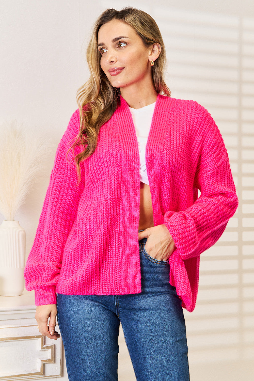 A woman wearing the Woven Right Rib-Knit Open Front Drop Shoulder Cardigan in bright pink over a white top and blue jeans stands indoors, looking to the side.