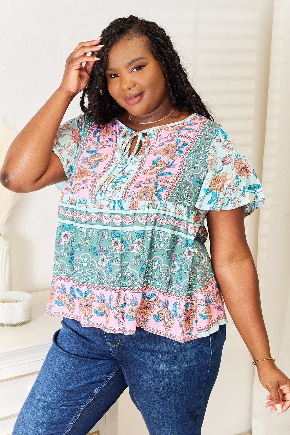 A person wearing a Perfee Floral Tie Neck Short Sleeve Blouse and denim jeans stands indoors, smiling slightly.