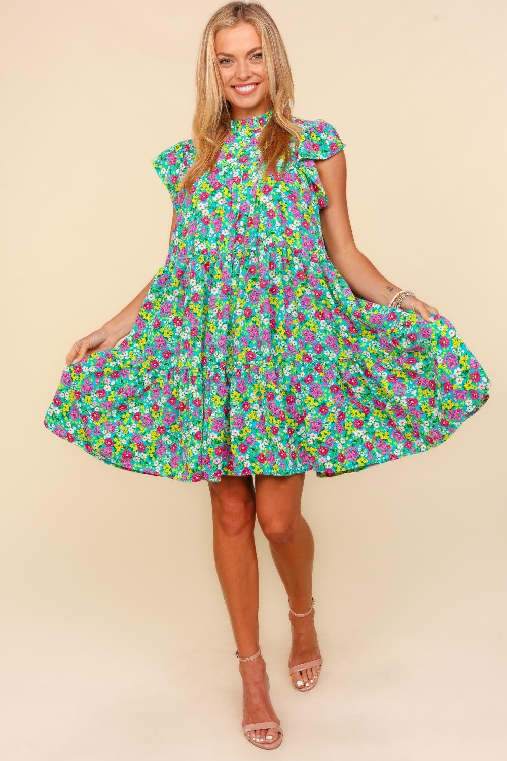 A woman in a Haptics Frilled Mock Neck Ditsy Floral Dress stands against a plain background, her beige high heels accentuating the feminine, elegant silhouette. Her hands rest casually in the pockets of her colorful, ditzy small floral print dress with flutter short sleeves.