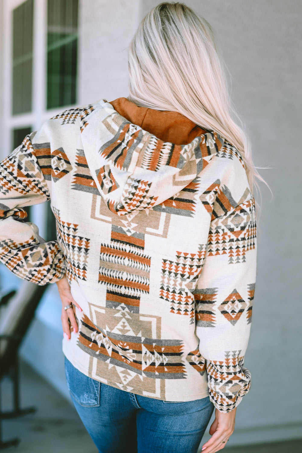 A person with long blonde hair, seen from behind, is wearing the Beige Aztec Print Kangaroo Pocket Half-Zip Hoodie. The hoodie showcases an Aztec pattern in shades of brown, beige, and orange and features a drop shoulder design with a cozy kangaroo pocket, all while standing outdoors.