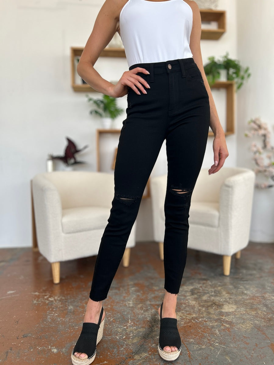 A person wearing a white T-shirt and Judy Blue Full Size Distressed Tummy Control High Waist Skinny Jeans poses barefoot in a room with white chairs and decorative plants in the background.