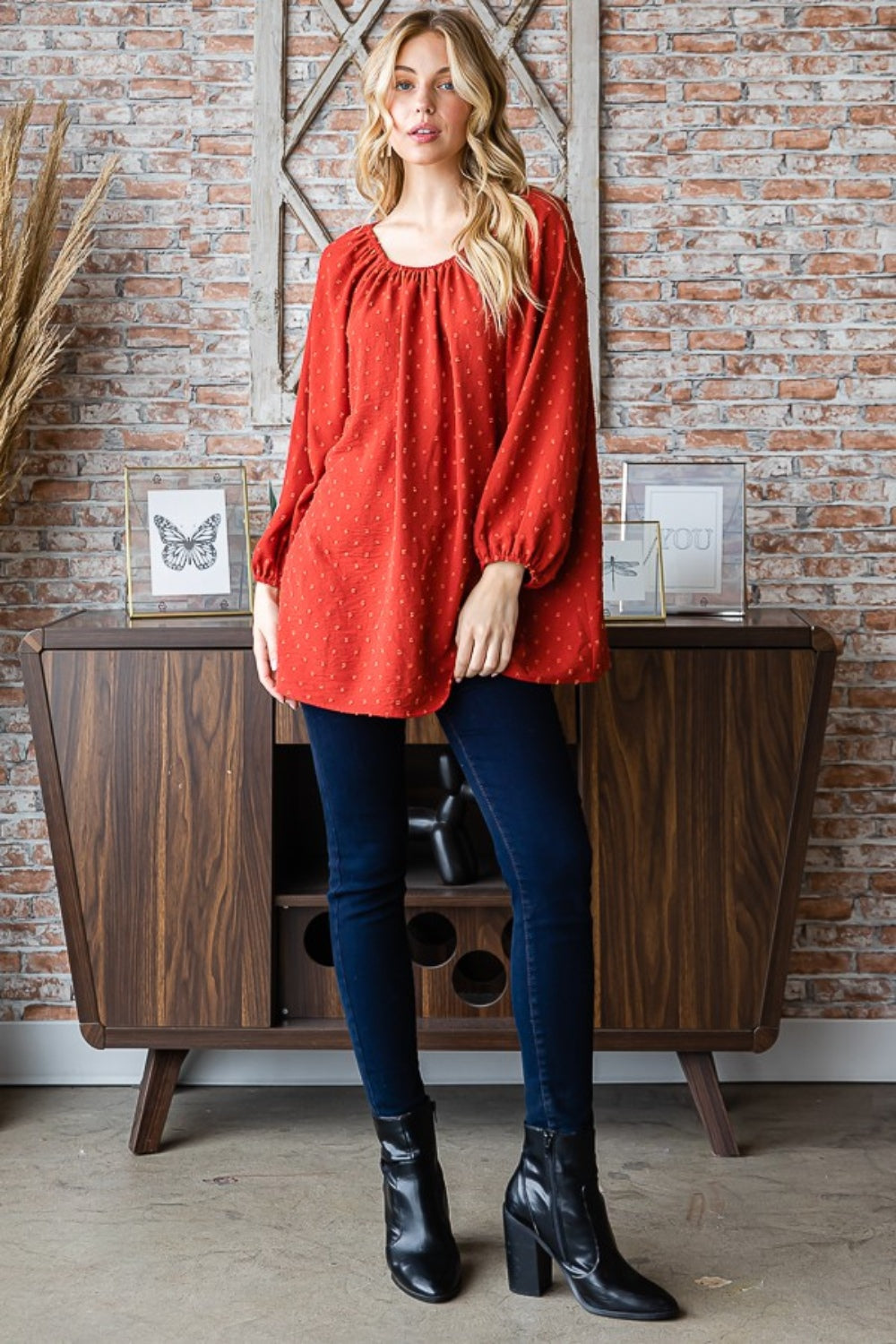 A woman wearing a Heimish Full Size Swiss Dot Off Shoulder Top paired with dark pants poses indoors against a brick wall decorated with butterfly artwork.