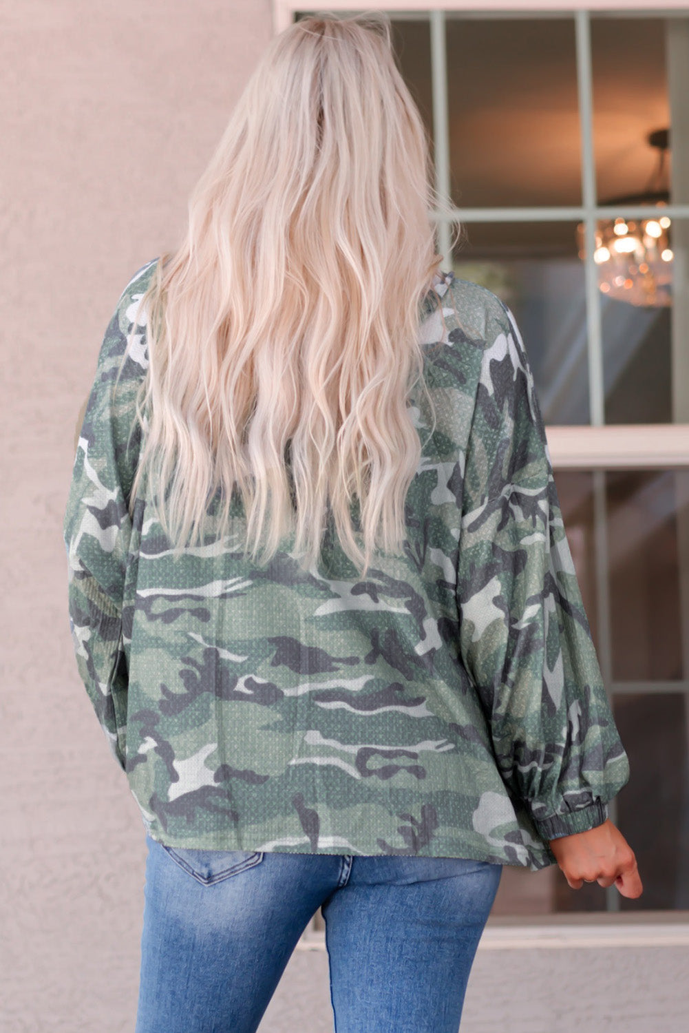 A person with long blonde hair is seen from behind, wearing a trendy Green Long Sleeve Camo Print Oversize Hoodie and blue jeans. They are standing in front of a window with a chandelier visible inside the building.