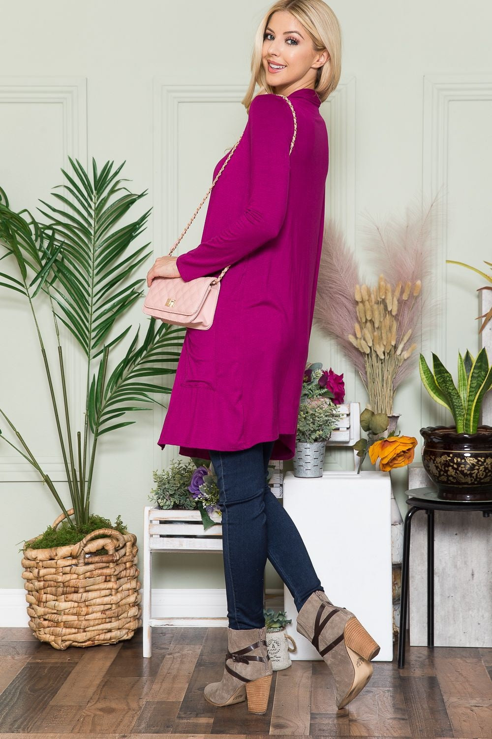 A woman with long blonde hair, wearing the Celeste Full Size Open Front Cardigan with Pockets in pink and a black top, poses with a light pink quilted purse on her shoulder. The open-front design of her versatile layering piece complements the indoor plants and light green wall behind her perfectly.