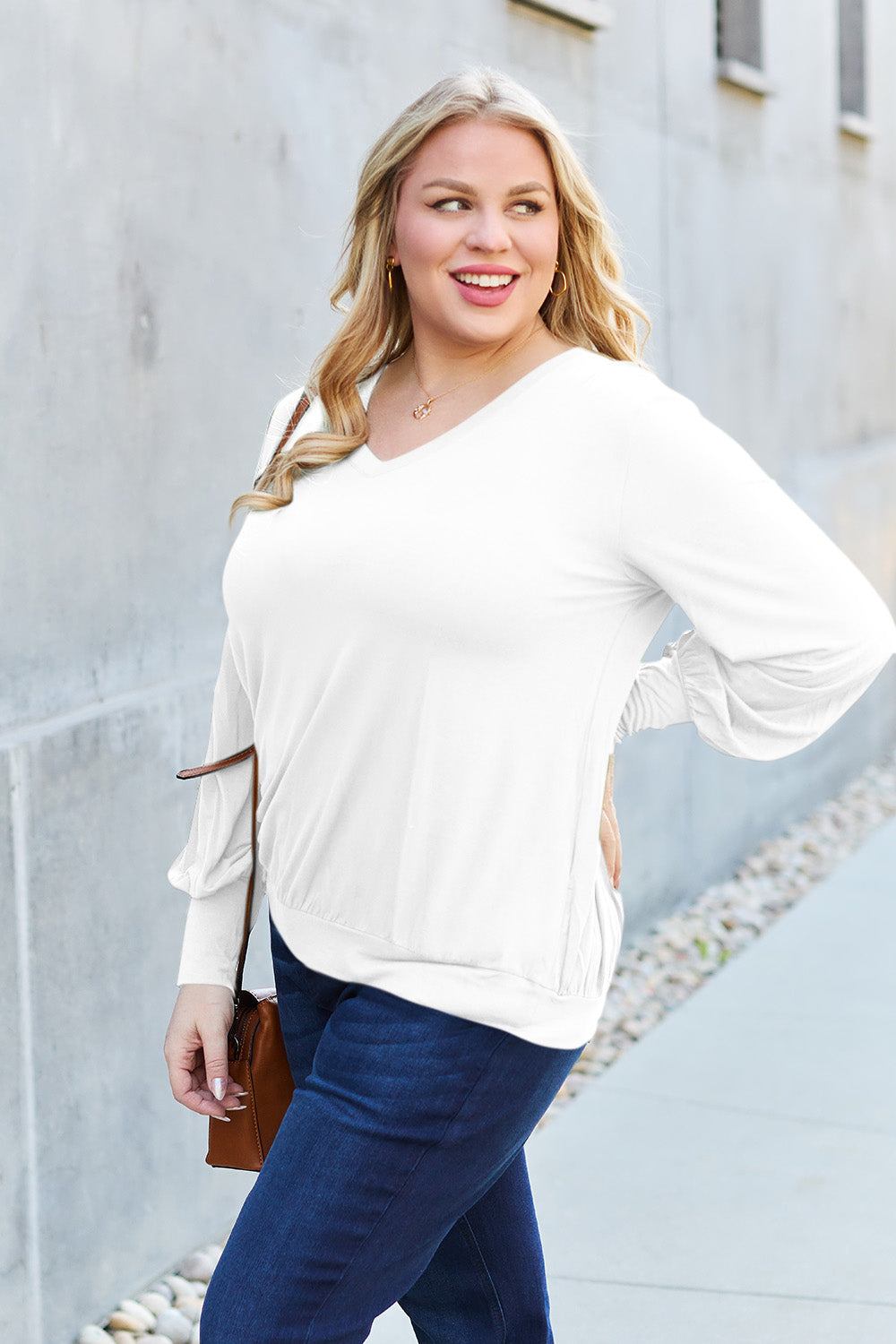 A woman with brown hair is standing against a concrete wall, wearing the Basic Bae Full Size V-Neck Lantern Sleeve Top in teal and blue basic-style jeans, smiling.