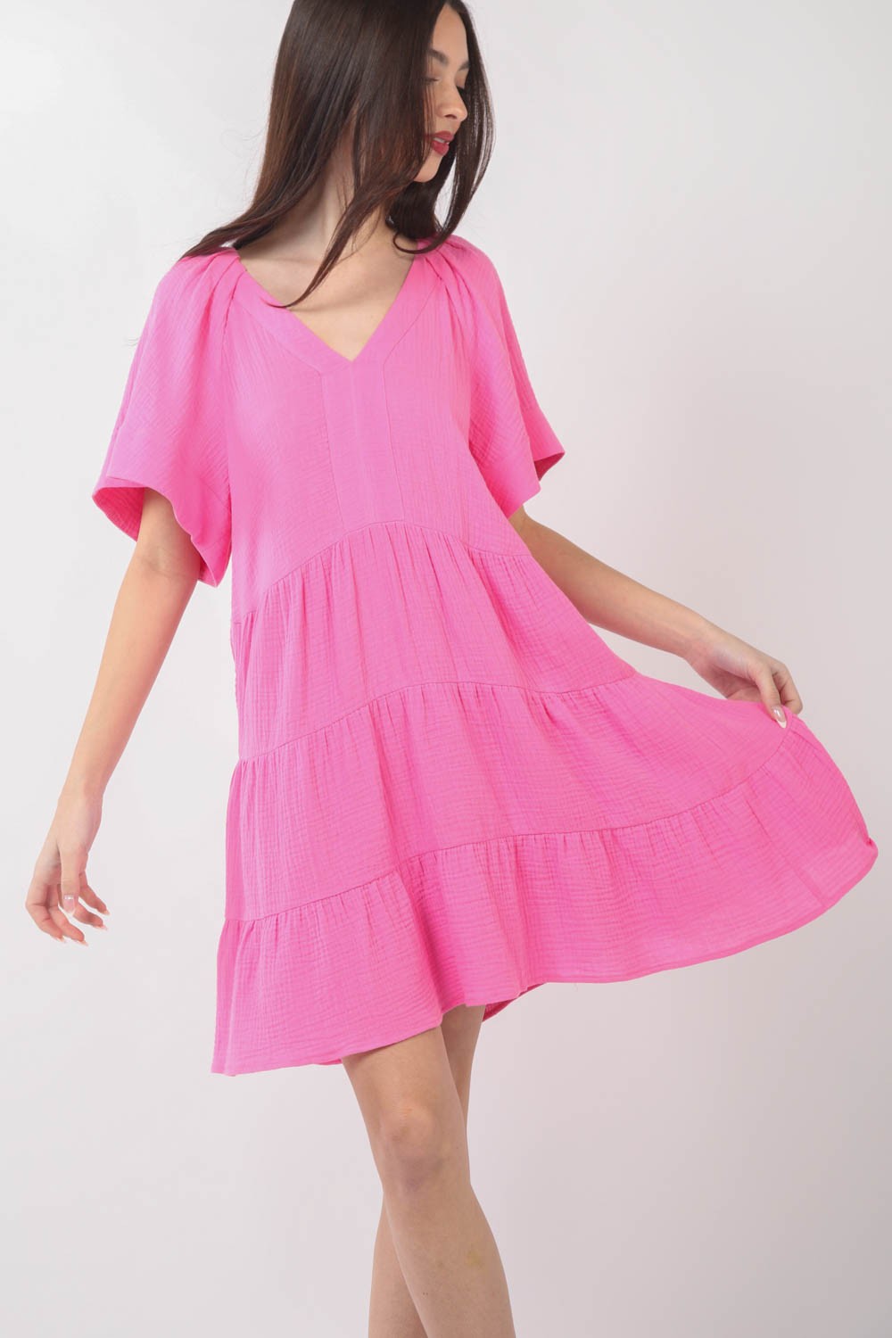 A woman is posing against a plain background, wearing the VERY J Texture V-Neck Ruffled Tiered Dress in pink, featuring short sleeves.