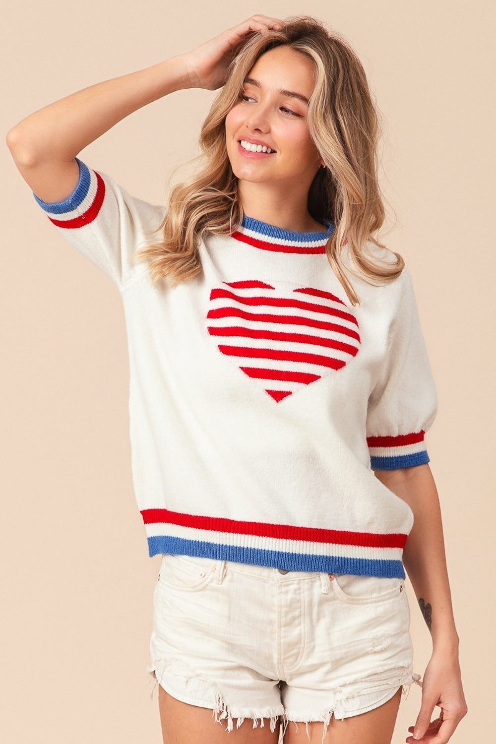 A person wearing the BiBi US Flag Theme Striped Heart Sweater, featuring a white short-sleeve design with red and blue stripes resembling the US flag and adorned with a red heart motif. They are also wearing white shorts and smiling with hands on hips.