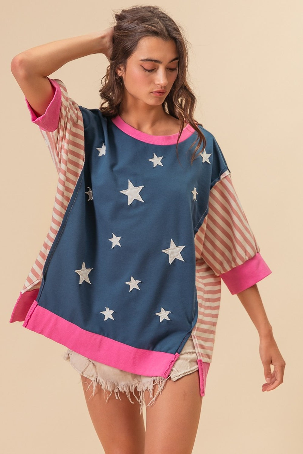 A person wearing a BiBi US Flag Theme Color Block Star Patch T-Shirt, characterized by its oversized fit, blue base with white star patterns, and pink-striped sleeves, stands against a plain background. They have their hand touching their hair.