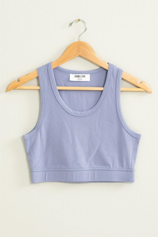 The "All I Need Cropped Tank Top," featuring a light blue hue and a fitted silhouette, hangs on a wooden hanger against a white wall.