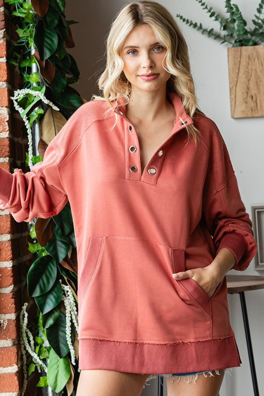 A woman with blonde hair is wearing a First Love Grommet Split Neck Kangaroo Pocket Sweatshirt in coral, standing indoors near a brick wall and plants.