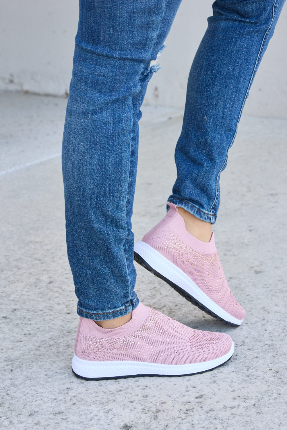 Person wearing Forever Link Rhinestone Mesh Breathable Sneakers in light pink and ripped blue jeans, standing on a concrete surface.