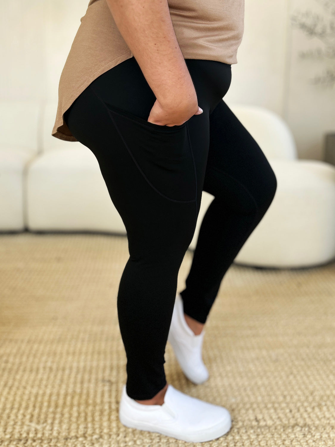 Person wearing Wide Waistband Sports Leggings made of stretchy nylon spandex and white slip-on shoes, standing on a beige carpet with a white couch in the background.