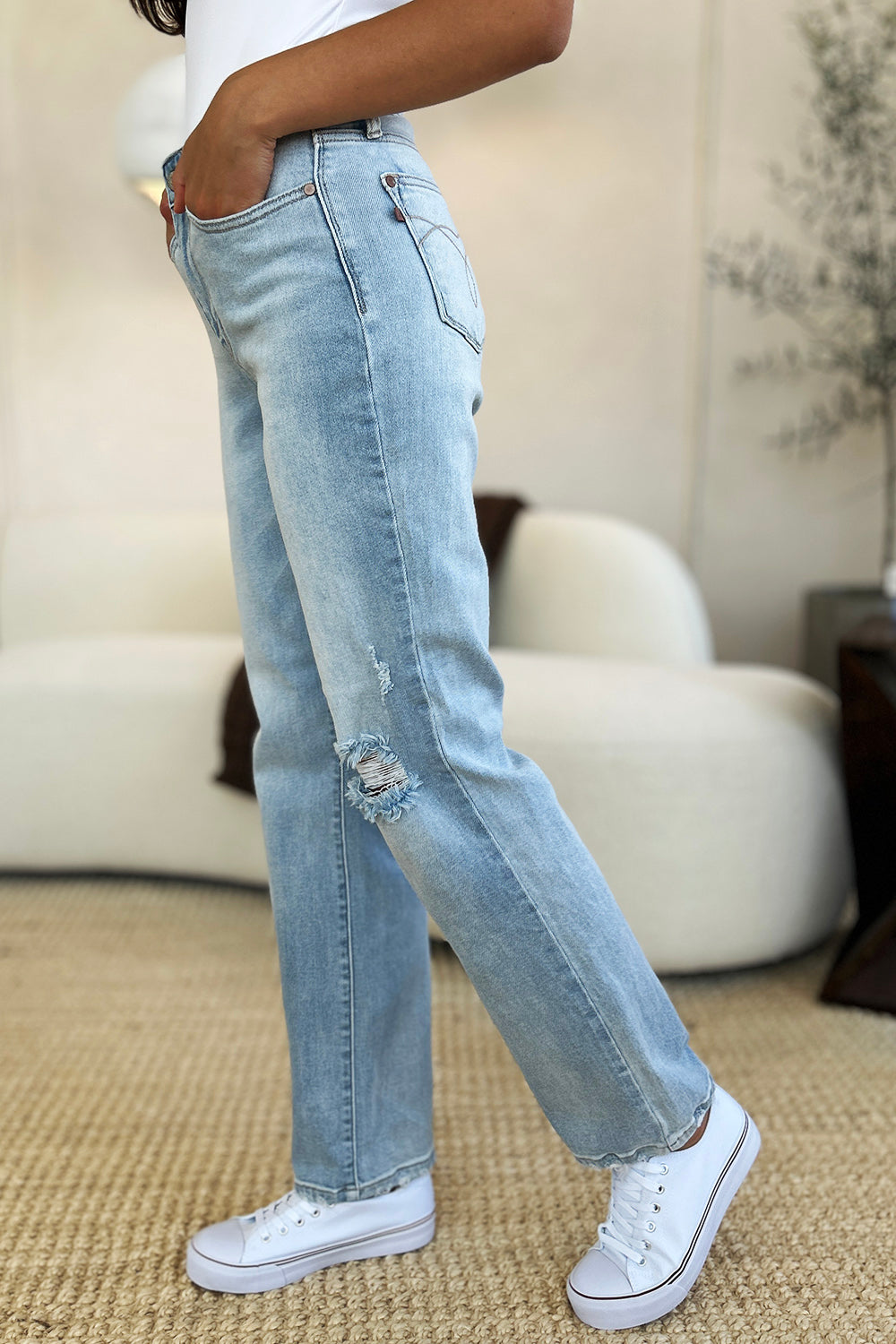 A person standing on a beige carpet, wearing Judy Blue Full Size High Waist Distressed Straight Jeans in light blue, a white top, and white sneakers.